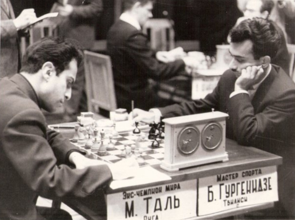 Mikhail Tal and Bukhuti Gurgenidze sit at a chess board with Baku chessmen, mid-game. Their table is labeled with their names in Cyrillic. An adjudicator attends.