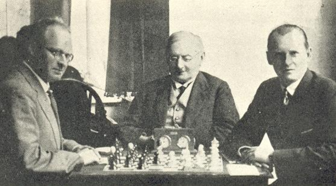 A black and white photo of Aron Nimzowitsch and Alexander Alekhine at a chess board, with Coffeehouse chessmen. Victor Tietz looks on.