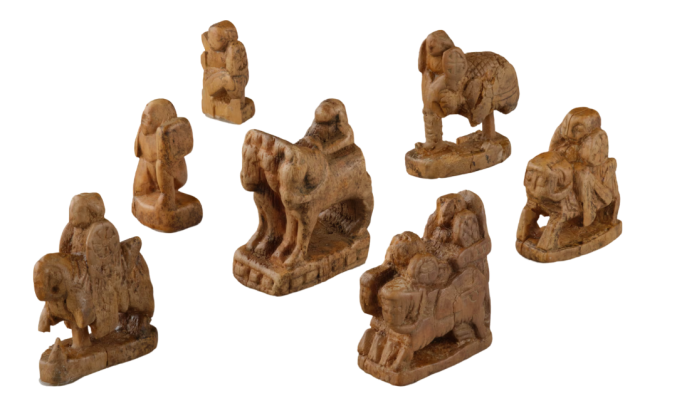 A photo of seven figurative chessmen stone chessmen. Two are charioteers, three are horsemen, and two are footsoldiers.